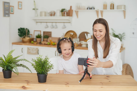 Close-up of a phone on a tripod on the kitchen table. Happy mom and daughter sit at the table in the kitchen and record educational videos for vloggingの写真素材