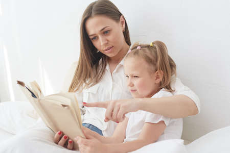 Young beautiful mother is reading a book to her daughter. Happy family, mother and daughter reading a book while lying in bed, smiling nanny mom telling funny story to cute preschool girlの写真素材