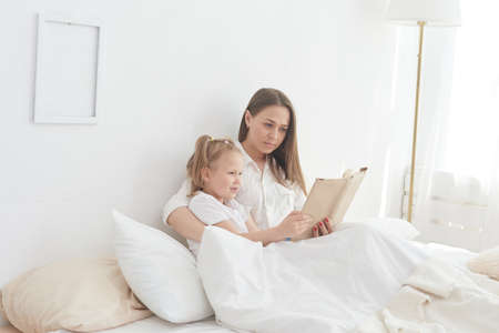 Close-up mom and little daughter are resting in a cozy bed, reading a funny interesting childrens book togetherの写真素材