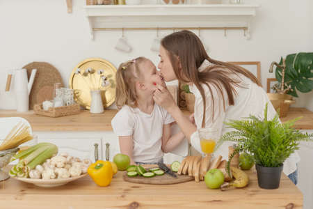 Cheerful mother and little daughter are preparing salad together in the kitchen and having fun. The girl kisses her mother at homeの写真素材