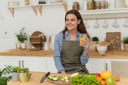 Beautiful woman preparing breakfast in a cozy wooden kitchen. A young woman is holding orange juice and is standing in her bright kitchenの写真素材