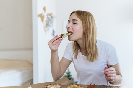 Happy young woman eating a piece of hot pizza at home and enjoying a delicious mealの写真素材