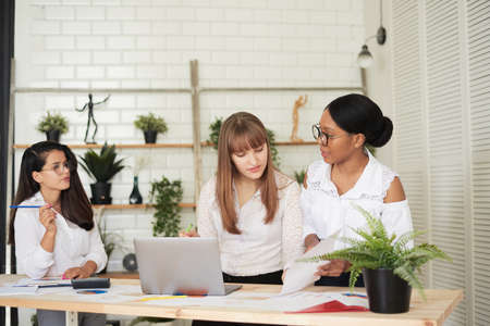 Serious female multiracial team solving problem together, diverse focused african, asian and caucasian colleagues discuss project brainstorming working on laptop, creative office teamworkの写真素材