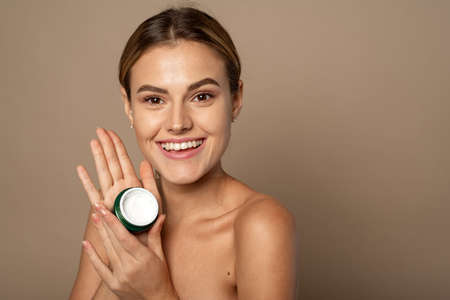 Portrait of a pretty young smiling woman with clean fresh skin in the studio on a beige background. Cute woman holding a jar of moisturizer in the studio. Skin care and hydration conceptの写真素材