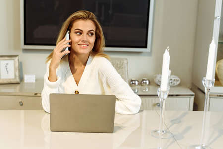 Young beautiful woman works for a computer from a home with a laptop on a white desk as a freelancer. Young businesswoman talking on the phone while working from homeの写真素材