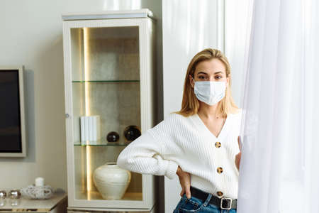 A young woman in a protective mask stands by the window. Self-isolation concept, protection against coronavirusの写真素材