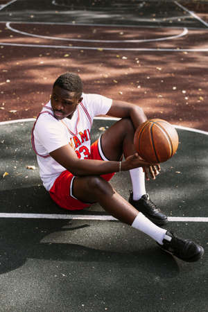 Portrait of an African American young man holding a basketball and sitting on a basketball court. Take a break during your workout. Fashionable sports portraitの写真素材