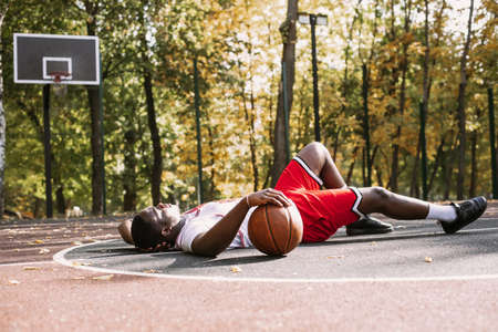 Portrait of an African American young man holding a basketball and sitting on a basketball court. Take a break during your workout. Fashionable sports portraitの写真素材