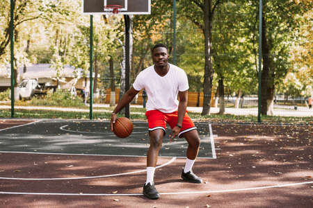 A young African American man is playing basketball on an outdoor court, smiling and looking at the camera. Outdoor sportsの写真素材