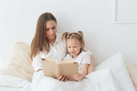 Young beautiful mother is reading a book to her daughter. Happy family, mother and daughter reading a book while lying in bed, smiling nanny mom telling funny story to cute preschool girlの写真素材