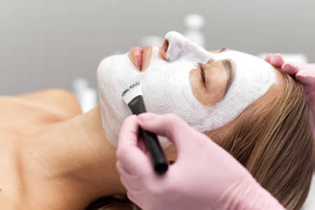 Beautician applies a white clay face mask with a brush on a woman's face. Professional beautician applies white cosmetic cream to a girl lying on a couch in a modern spa. Close up. Relaxed woman enjoying wellness facials.の写真素材