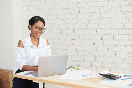 Portrait of a handsome smiling African American office worker sitting at a desk using a computer. Young african american businesswoman working on laptop in modern officeの写真素材