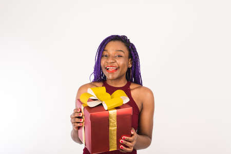 A black woman holds a huge red gift with a gold bow in her hands, looks at the camera and smiles. Concept of holiday, New year and Christmasの写真素材
