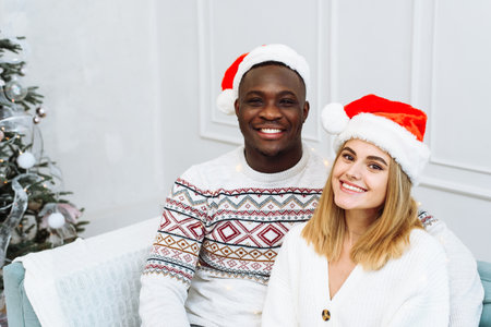 A young multiethnic couple in santa hats are sitting on a sofa in a bright interior next to a tree. Man and woman looking at the camera, smilingの写真素材