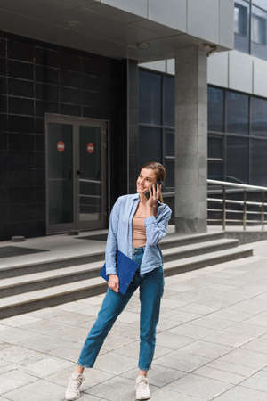 A businesswoman stands against the background of the office center, smiles and speaks on the phone. Communication with business partners at a distance. Young Female Business Styleの写真素材