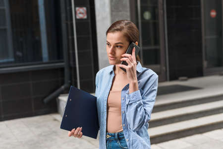 A businesswoman stands against the background of the office center, smiles and speaks on the phone. Communication with business partners at a distance. Young Female Business Styleの写真素材