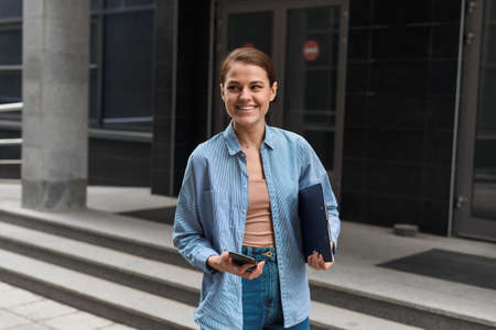 A businesswoman stands against the background of the office center, holds a phone in her hands and looks at the camera, smilingの写真素材