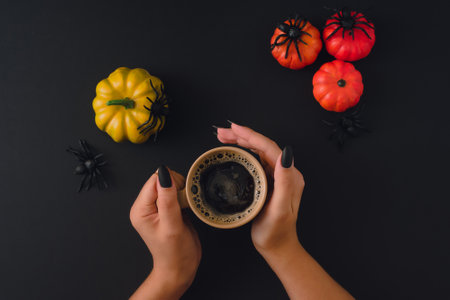A female hand holds a mug with coffee on a black background with decorative spiders and pumpkins. Traditional, party and festive concept. Halloween. flat top viewの写真素材