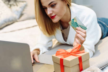 Happy young woman with red lipstick lies on the bed and buys Christmas gifts for her friends using a credit card. Buying Christmas and birthday gifts. Holiday Salesの写真素材