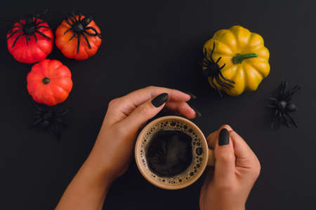 Black manicured hands holding cappuccino mug on Halloween background with devotive pumpkins and spiders. View from aboveの写真素材