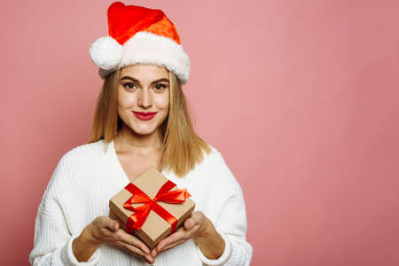 Lovely young woman in santa hat is holding a Christmas present, smiling and looking at the camera. Beautiful blonde on a pink background in the studio. Concept for Christmas shopping, gift giving and New Years.の写真素材