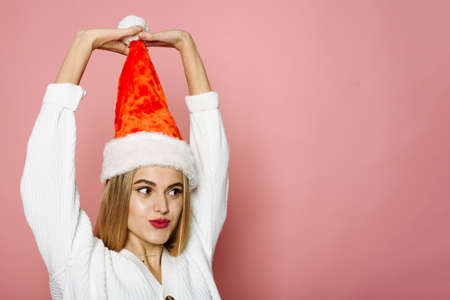 Funny woman holding a pompom of santa hat and looking to the camera on a pink background. Smiling happy womanの写真素材