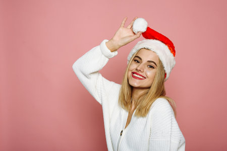 Funny woman holding a pompom of santa hat and looking to the camera on a pink background. Smiling happy womanの写真素材