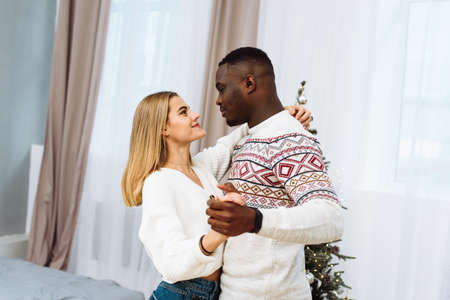 A young multi-ethnic couple is enjoying each other in a dance next to the Christmas tree. Cozy moments of Christmas daysの写真素材