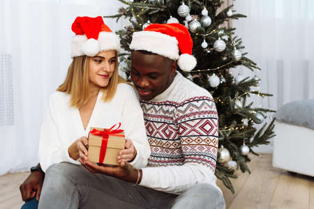 Joyful cozy moments in winter holidays. A multinational couple opens a Christmas present. A loving young black man and a white woman are sitting next to the Christmas treeの写真素材