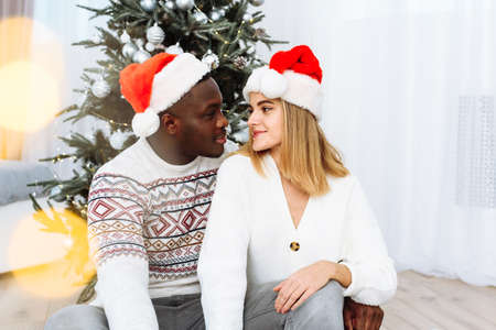 Portrait of a couple in love in santa hats sitting by the tree. Glare of the yellow garland. Black male hugs a Caucasian womanの写真素材