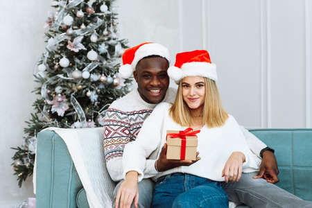 Couple sharing a Christmas present. Portrait of a lovely couple during Christmas time. Young happy multiethnic couple hugging and relaxing on the comfortable sofa. Young man and woman are looking at the camera and are smiling with all their teeth.の写真素材