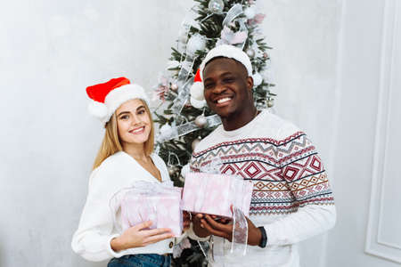 Young multinational couple in Santa hats hold gifts against the background of a Christmas tree and smile while looking at the camera. Christmas and New Years conceptの写真素材
