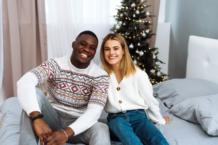 A couple in love sits next to each other on the bed against the background of a Christmas tree and smile at the camera. Christmas and New Year conceptの写真素材