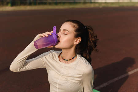 Young beautiful sports model drinks water during a break at the outdoor stadium in the parkの写真素材