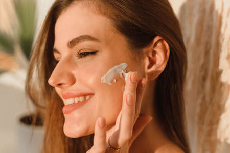 A young woman applies moisturizer to her face in her sun bath. Daily routine, hydration and skin care conceptの写真素材