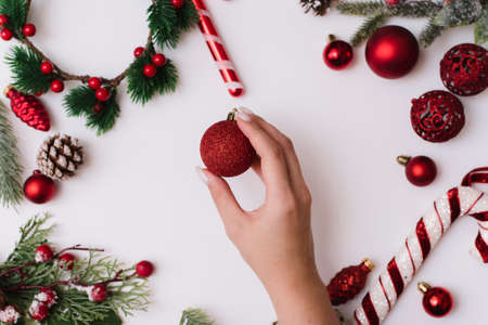 A female hand with a white manicure holds a red ball on a white background with Christmas branches and red decorationsの写真素材