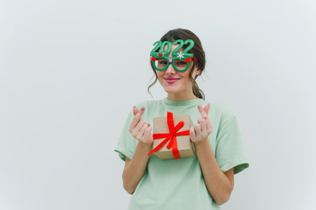 Modern young woman wearing 2022 glasses is holding a Christmas present and showing a Korean heart sign.の写真素材