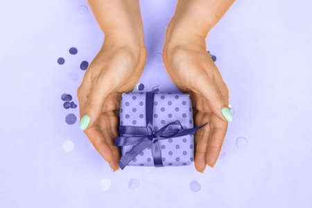 Womans hands with blue pastel manicure holding a gift with a bow on a purple background very peri 2022. The concept of the holiday, gifts, surprises, birthday, new year and Christmasの写真素材