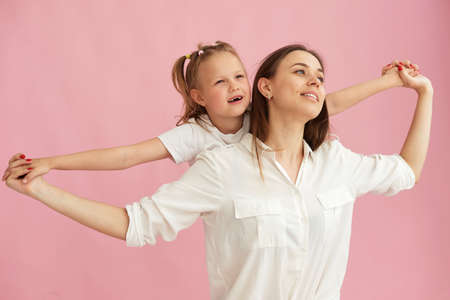 Mom woman in light clothes has fun with cute baby girl. Little baby daughter sitting on mothers back isolated on pastel pink background. studio portraitの写真素材
