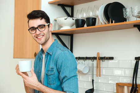 A handsome, positive, smiling, young guy holds a cup of tea or coffee in his hands, looks out the window, stands in the kitchenの写真素材