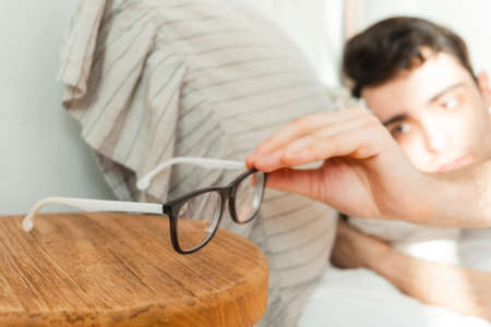 A young man reaches for his glasses after waking up from a dream in bed.の写真素材