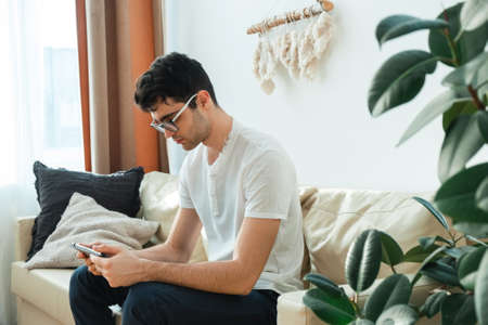 Attractive young man is using a tablet while sitting on the couch at home and browsing social networks and news.の写真素材