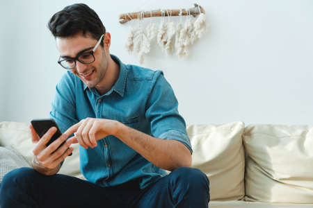 Portrait of an attractive smiling young man in casual clothes sitting on the sofa in the living room and watching the news using a mobile phone.の写真素材
