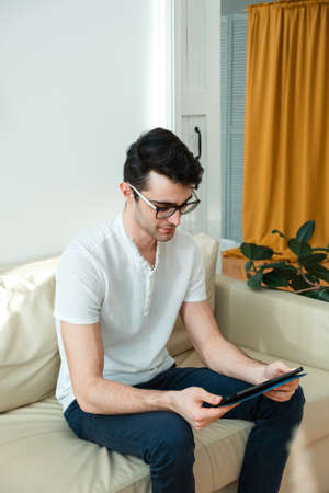 Young man browsing internet or checking social media on tablet, relaxing on sofa at home.の写真素材