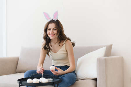 Beautiful young woman in bunny ears paints eggs for easter and looks at the camera.の写真素材