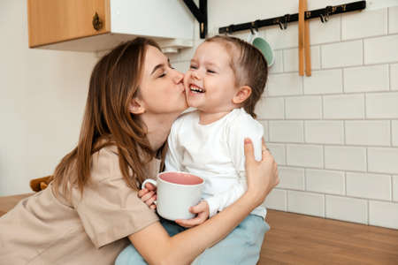 Loving mom kissing her little daughter in the kitchen, cheerful family posing while having breakfast at home together, having fun and enjoying the home atmosphere, close-up. Mothers Dayの写真素材