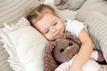 Beautiful little girl hugging a plush bunny while sleeping in bed at home. Peaceful baby sleepの写真素材