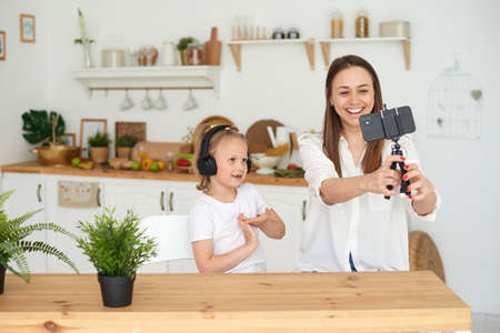 A happy family. Cute little daughter is sitting next to mom in the kitchen, laughing, having fun using a smartphone, watching a funny video. Video communication during quarantine, communication with relatives and friends using a smartphone.の写真素材