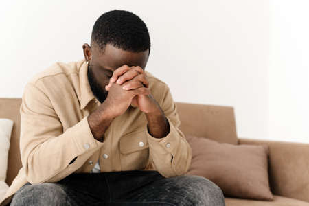 African American believing man sitting at home on sofa praying to God, asking for good luck, thoughtful bi-racial superstitious man holding hands in prayer, feeling gratitude, faith, religion conceptの写真素材