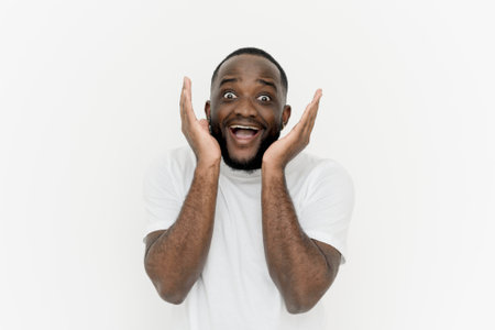 My God! Amazed african american man screaming excitedly and looking at camera on white background. A black young man gesticulates with his hands, expresses positivity and joy after hearing the good news.の写真素材
